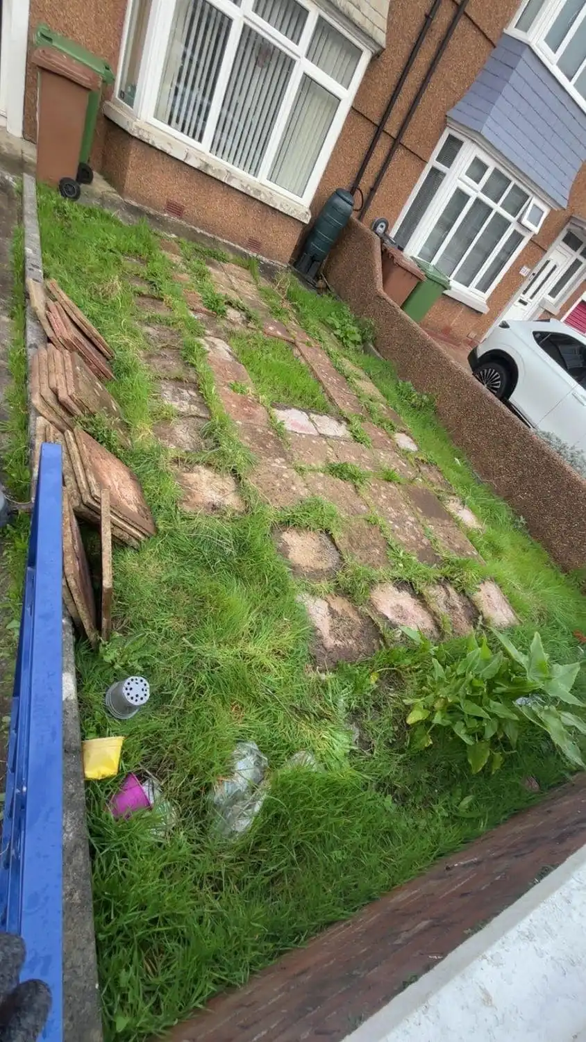 A front garden of broken, uneven slabs with grass and weeds growing through every joint