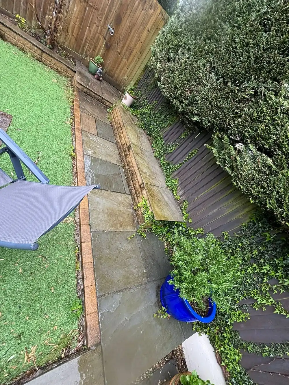 A narrow garden path beside dark decking, with an overgrown border spilling across the slabs