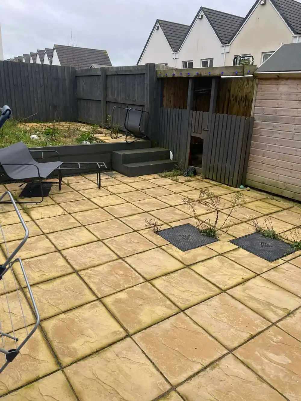 A large buff-coloured patio with weeds and grass growing up through the joints between the slabs