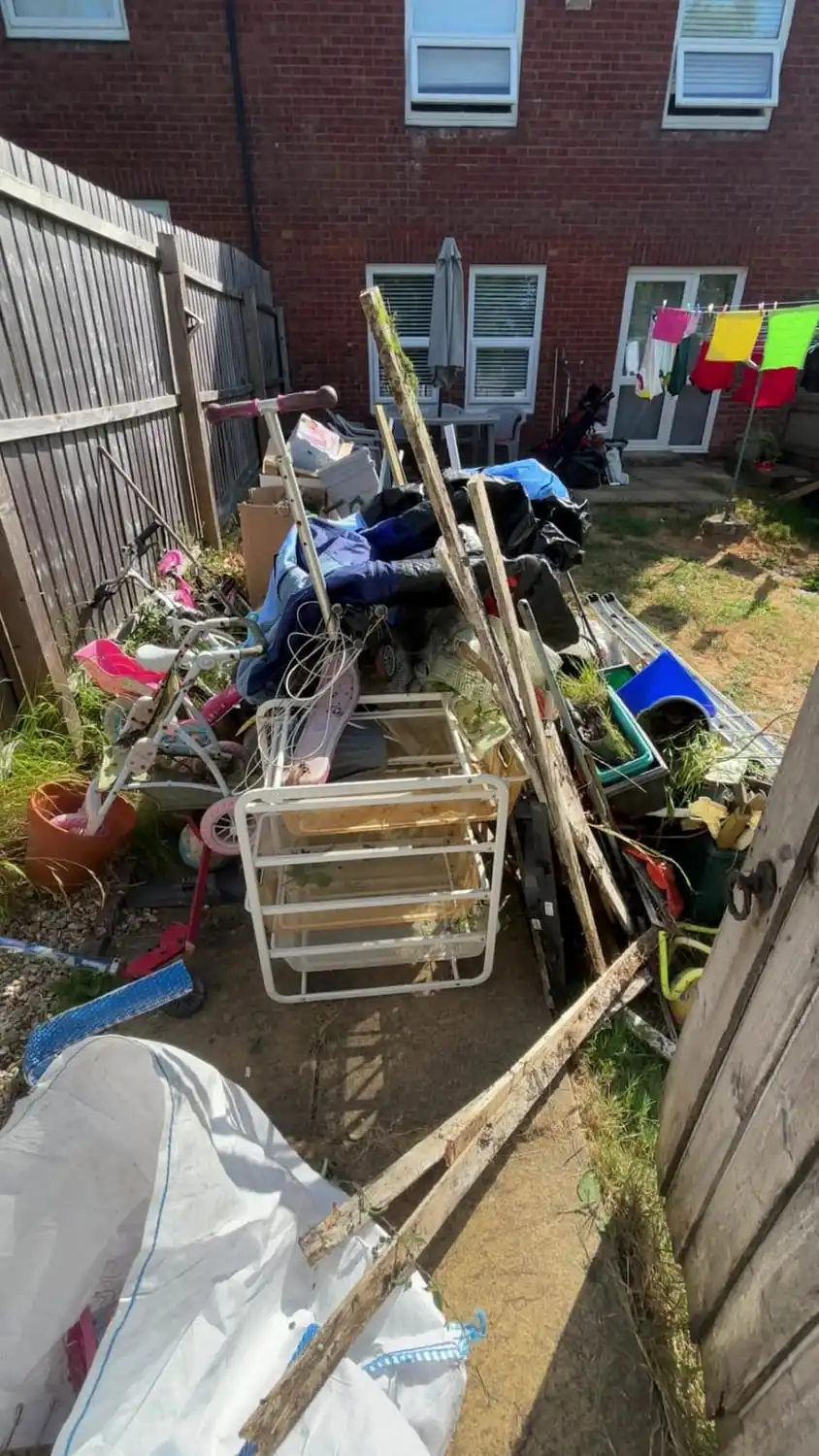 A back yard piled with old garden furniture, bicycles, pots and household items stacked against the fence